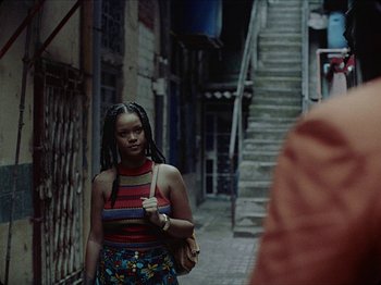 Movie still from “Guava Island” (2019), directed by Hiro Murai – A young woman walking down a street with a purse; Medium shot, Over the shoulder angle