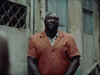Movie still from “Guava Island” (2019), directed by Hiro Murai – A man in an orange shirt standing in front of a building; Medium shot, Low angle