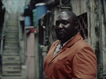 Movie still from “Guava Island” (2019), directed by Hiro Murai – A man in a red jacket standing in a street; Medium shot, Low angle