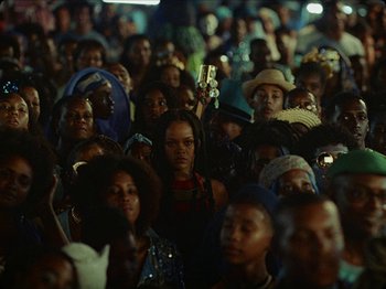 Movie still from “Guava Island” (2019), directed by Hiro Murai – A group of people gathered together in a crowd; Medium shot, High angle
