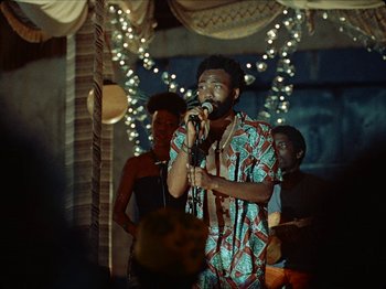 Movie still from “Guava Island” (2019), directed by Hiro Murai – A man is singing into a microphone in front of a group of people; Medium shot, Over the shoulder angle