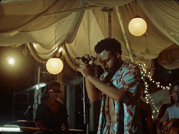 Movie still from “Guava Island” (2019), directed by Hiro Murai – A man with a microphone in front of a crowd; Medium shot, Over the shoulder angle
