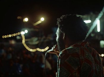 Movie still from “Guava Island” (2019), directed by Hiro Murai – A man standing in front of a microphone on a stage; Medium shot, Low angle