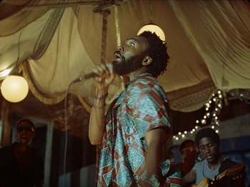Movie still from “Guava Island” (2019), directed by Hiro Murai – A man with a beard is singing into a microphone; Medium shot, Low angle
