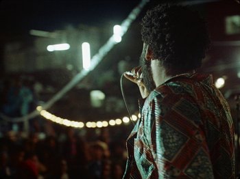Movie still from “Guava Island” (2019), directed by Hiro Murai – A man with curly hair is holding a microphone; Medium shot, Low angle