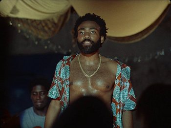 Movie still from “Guava Island” (2019), directed by Hiro Murai – A man with a beard is standing in front of a crowd; Medium shot, Low angle