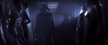 Movie still from “I Know What You Did Last Summer” (1997), directed by Jim Gillespie – A man in a black coat and a black hat is standing in a dark room; Wide shot, Low angle