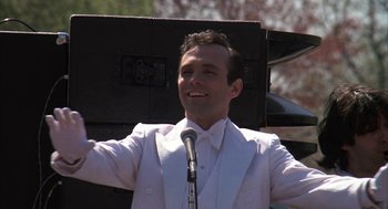 Movie still from “Hair” (1979), directed by Milos Forman – A man in a white suit is holding a microphone; Medium shot, Low angle