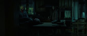 Movie still from “Halloween Ends” (2022), directed by David Gordon Green – Two people in a dark room with a table and a window in the background; Wide shot, Over the shoulder angle