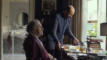 Movie still from “Happy End” (2017), directed by Michael Haneke – A man standing at a table with a man cutting a cake; Medium shot, Over the shoulder angle