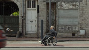 Movie still from “Happy End” (2017), directed by Michael Haneke – An older man in a suit is sitting in a wheel chair on the sidewalk; Wide shot, Low angle
