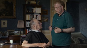 Movie still from “Happy End” (2017), directed by Michael Haneke – A man standing next to an older man in front of a book shelf; Medium shot, Over the shoulder angle
