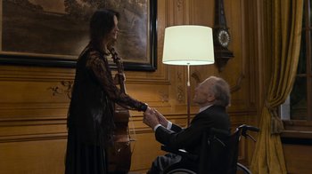 Movie still from “Happy End” (2017), directed by Michael Haneke – An older man in a wheelchair shaking hands with an older woman in a black dress; Medium shot, Over the shoulder angle