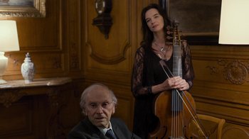 Movie still from “Happy End” (2017), directed by Michael Haneke – An older man sitting next to a woman holding a cello; Medium shot, Over the shoulder angle