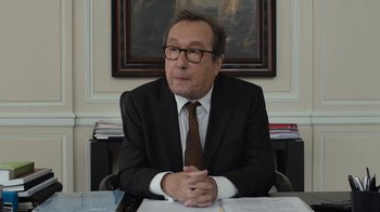 Movie still from “Happy End” (2017), directed by Michael Haneke – An older man sitting at a desk in a suit and tie; Close Up shot, Over the shoulder angle
