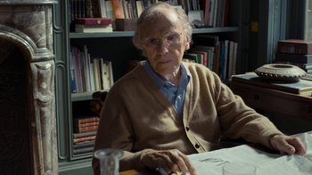 Movie still from “Happy End” (2017), directed by Michael Haneke – An older man sitting at a table in front of a book shelf; Close Up shot, Over the shoulder angle