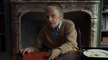 Movie still from “Happy End” (2017), directed by Michael Haneke – An older woman sitting at a table in front of a fire place; Medium shot, High angle