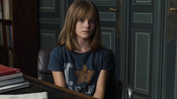Movie still from “Happy End” (2017), directed by Michael Haneke – A young girl sitting in a chair in front of a door; Medium shot, High angle