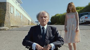Movie still from “Happy End” (2017), directed by Michael Haneke – An old man in a wheel chair and a young girl in a dress; Medium shot, Low angle
