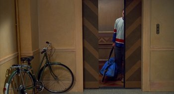 Movie still from “Happy Gilmore” (1996), directed by Dennis Dugan – A man holding a bag and a bicycle in a room; Wide shot, Overhead angle