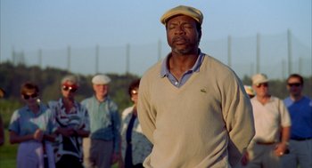 Movie still from “Happy Gilmore” (1996), directed by Dennis Dugan – A man in a tan sweater and a hat is standing in front of a group of people; Close Up shot, Low angle