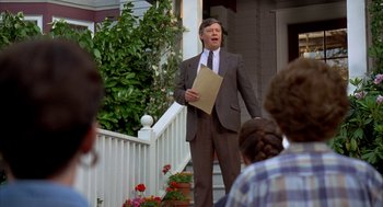 Movie still from “Happy Gilmore” (1996), directed by Dennis Dugan – A man in a brown suit holding a piece of paper while standing in front of a house; Medium shot, Over the shoulder angle