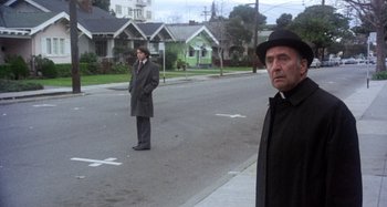 Movie still from “Harold and Maude” (1971), directed by Hal Ashby – A man in a black hat standing on the side of the street; Medium shot, Over the shoulder angle