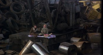 Movie still from “Harold and Maude” (1971), directed by Hal Ashby – Two women sitting on a blanket in a warehouse; Wide shot, High angle