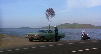 Movie still from “Harold and Maude” (1971), directed by Hal Ashby – A man standing next to a car on the side of the road; Wide shot, High angle