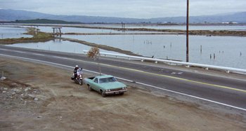 Movie still from “Harold and Maude” (1971), directed by Hal Ashby – A man riding a motorcycle next to a car on the side of a road; Extreme Wide shot, High angle