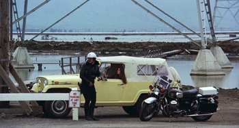 Movie still from “Harold and Maude” (1971), directed by Hal Ashby – A man standing next to a yellow car and a motorcycle; Wide shot, High angle