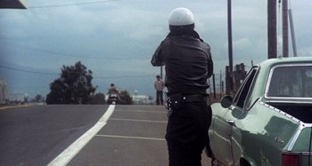 Movie still from “Harold and Maude” (1971), directed by Hal Ashby – A man in a motorcycle helmet is standing on the side of the road; Wide shot, Low angle