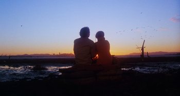 Movie still from “Harold and Maude” (1971), directed by Hal Ashby – Two people sitting on a rock at the beach at sunset; Wide shot, Low angle