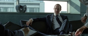 Movie still from “Headhunters” (2011), directed by Morten Tyldum – A man in a suit and tie sitting on a couch; Medium shot, Over the shoulder angle