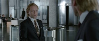 Movie still from “Headhunters” (2011), directed by Morten Tyldum – A man in a suit and tie standing in front of a building; Medium shot, Over the shoulder angle