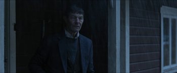 Movie still from “Headhunters” (2011), directed by Morten Tyldum – An older man wearing a suit standing in front of a window; Medium shot, Over the shoulder angle