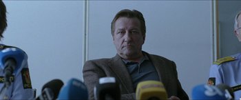 Movie still from “Headhunters” (2011), directed by Morten Tyldum – A man sitting in front of microphones in front of a wall; Medium shot, Over the shoulder angle