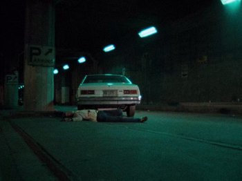 Movie still from “Henry: Portrait of a Serial Killer” (1986), directed by John McNaughton – A man laying on the ground next to a car; Wide shot, High angle