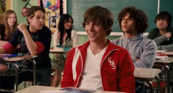 Movie still from “High School Musical 3: Senior Year” (2008), directed by Kenny Ortega – A man sitting at a table in front of a group of people; Medium shot, Over the shoulder angle