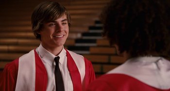 Movie still from “High School Musical 3: Senior Year” (2008), directed by Kenny Ortega – A young man in a graduation gown talking to another young man; Medium shot, Over the shoulder angle