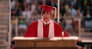 Movie still from “High School Musical 3: Senior Year” (2008), directed by Kenny Ortega – A boy in a red cap and gown at a graduation ceremony; Medium shot, Low angle