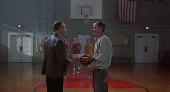 Movie still from “Hoosiers” (1986), directed by David Anspaugh – Two men standing next to each other holding a basketball in a gym; Wide shot, Over the shoulder angle