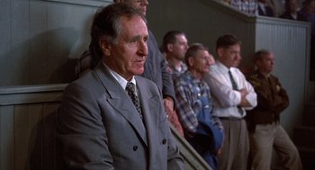Movie still from “Hoosiers” (1986), directed by David Anspaugh – A man in a suit and tie sitting in front of a group of men; Medium shot, Low angle