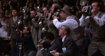 Movie still from “Hoosiers” (1986), directed by David Anspaugh – A group of men in suits taking pictures in a crowd; Wide shot, Low angle