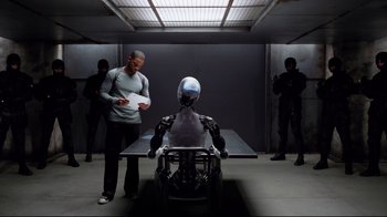 Movie still from “I, Robot” (2004), directed by Alex Proyas – A man standing in front of a table with a robot on top of it; Wide shot, High angle