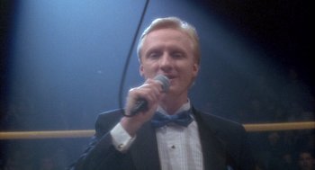 Movie still from “Hot Shots!” (1991), directed by Jim Abrahams – A man in a suit and bow tie holding a microphone; Close Up shot, Low angle