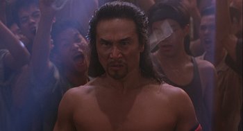 Movie still from “Hot Shots! Part Deux” (1993), directed by Jim Abrahams – A man with long black hair and no shirt in front of a group of people; Close Up shot, Low angle