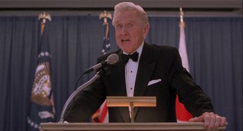 Movie still from “Hot Shots! Part Deux” (1993), directed by Jim Abrahams – An older man in a tuxedo is speaking at a podium; Close Up shot, Low angle