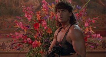 Movie still from “Hot Shots! Part Deux” (1993), directed by Jim Abrahams – A man in a tank top holding a gun in front of a bunch of flowers; Medium shot, Low angle