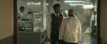 Movie still from “Hotel Mumbai” (2018), directed by Anthony Maras – A man wearing a turban talking to another man in a kitchen; Medium shot, Over the shoulder angle